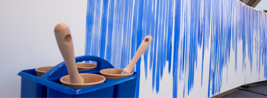 5 of 8, Blue paint bucket with brushes in front of white canvases with blue lines